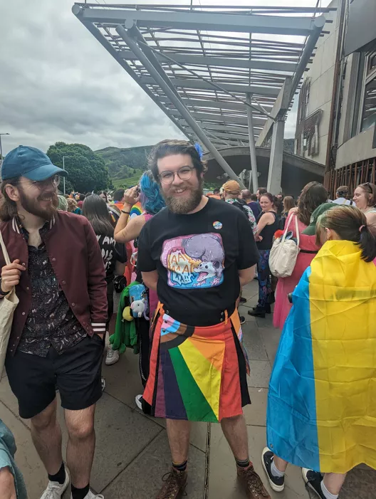 Me wearing a pride flag kilt, black shirt and smiling holding a champagne glass