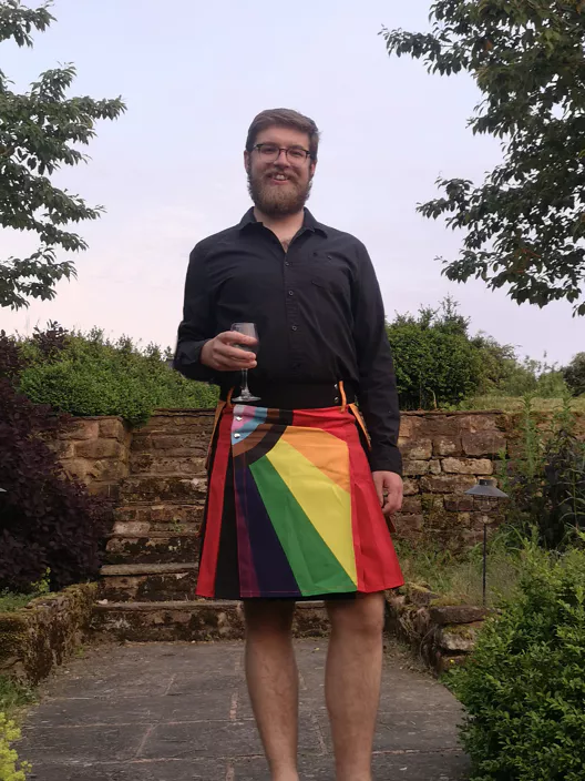 Me wearing a pride flag kilt, black shirt and smiling holding a champagne glass